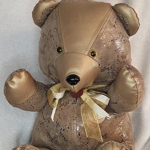 May include: A plush teddy bear with a gold and brown color scheme. The bear has a textured body, gold accents on the paws, ears, and head, and a bow around its neck. A small tag with the word "SYLVAN" hangs from its foot.
