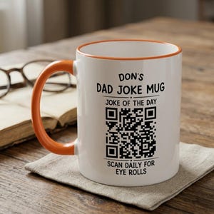 May include: A white ceramic mug with an orange handle and rim. The front features black text that reads DON'S DAD JOKE MUG, JOKE OF THE DAY, and SCAN DAILY FOR EYE ROLLS, surrounding a central black and white QR code for scanning purposes.