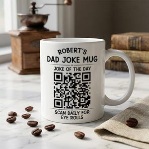May include: A white ceramic mug featuring black text that reads ROBERT'S DAD JOKE MUG, JOKE OF THE DAY, and SCAN DAILY FOR EYE ROLLS, centered around a large black and white QR code. The mug has a standard handle and a smooth, cylindrical body.