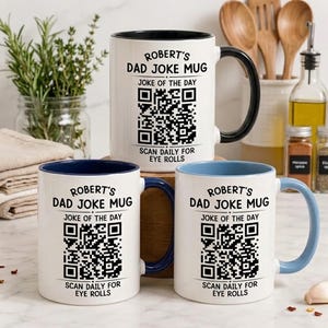 May include: Three white ceramic mugs with black, dark blue, and light blue interiors and handles. Each mug features the text 'ROBERT'S DAD JOKE MUG', 'JOKE OF THE DAY', and 'SCAN DAILY FOR EYE ROLLS' surrounding a black and white QR code.