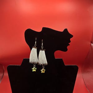 May include: A pair of dangle earrings displayed on a black jewelry stand. Each earring features a white, fringed design with a brown accent and a dangling star-shaped charm. The background is a vibrant red.