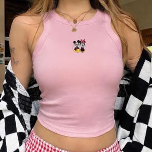 Puede incluir: Camiseta sin mangas rosa claro con una peque&ntilde;a imagen bordada de Mickey y Minnie Mouse. La camiseta tiene una textura acanalada y un cuello redondo. Los personajes de dibujos animados visten sus trajes cl&aacute;sicos. La camiseta es sin mangas.