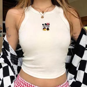 Puede incluir: Una camiseta corta blanca sin mangas con una peque&ntilde;a imagen bordada de Mickey y Minnie Mouse. La parte superior es de color s&oacute;lido y tiene un cuello redondo. La modelo lleva una chaqueta a cuadros en blanco y negro.