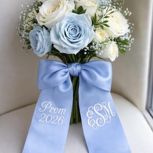May include: A bouquet of white and light blue roses with baby's breath. The flowers are tied with a light blue satin ribbon, featuring the text "Prom 2026" and initials in white script.