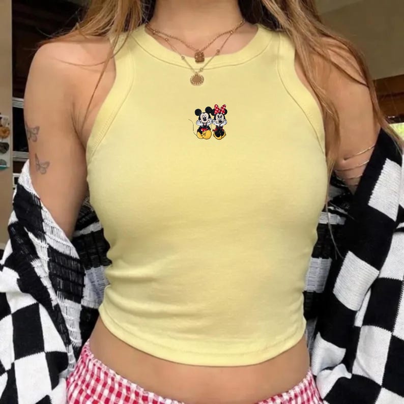 Puede incluir: Top corto amarillo claro con una peque&ntilde;a imagen bordada de Mickey y Minnie Mouse. El top es sin mangas y tiene un cuello redondo. La persona lleva una chaqueta a cuadros blancos y negros.