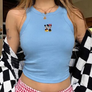 Puede incluir: Una camiseta sin mangas azul claro con textura acanalada. La parte superior presenta una imagen bordada de Mickey y Minnie Mouse. La modelo lleva una rebeca a cuadros en blanco y negro y collares dorados.