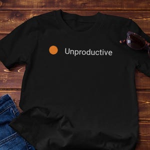 May include: Black t-shirt with the word "Unproductive" and an orange dot. The shirt is paired with blue jeans and sunglasses on a wooden surface. A small succulent plant in a white pot is also present.