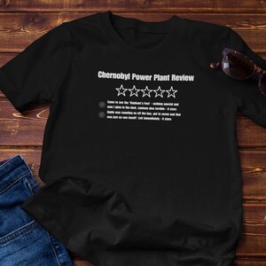 Könnte beinhalten: Schwarzes T-Shirt mit dem Text "Chernobyl Power Plant Review" und einem Bewertungssystem. Das Shirt liegt auf einer Holzoberfläche mit einer Jeans, einer Sonnenbrille und einer kleinen Topf-Sukkulente. Der Text enthält eine Bewertung.