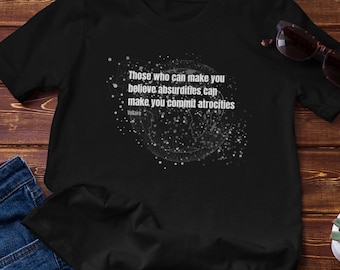 May include: Black t-shirt featuring the quote "Those who can make you believe absurdities can make you commit atrocities" in white. The image includes blue jeans and sunglasses, set on a wooden surface.