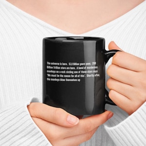 May include: Black ceramic mug with a white, humorous quote about the universe and monkeys. The mug is held by a person wearing a white sweater. The text on the mug reads: "The universe is born...the monkeys blow themselves up."