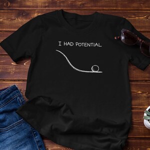 Könnte beinhalten: Ein schwarzes T-Shirt mit kurzen Ärmeln und einer weißen Grafik, die einen Ball am unteren Ende einer geschwungenen Linie zeigt, darüber der Text I HAD POTENTIAL. Das T-Shirt liegt neben einer blauen Jeans, einer Sonnenbrille und einer kleinen Sukkulente.