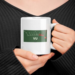 May include: White ceramic mug featuring a green VU meter graphic with the text "VU". The meter needle is red. The mug is held by a person wearing a black sweater.