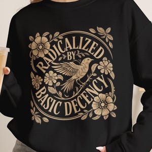 Puede incluir: Una sudadera negra de manga larga con un estampado gráfico en color tostado de un pájaro rodeado de flores y el texto "RADICALIZED BY BASIC DECENCY" dispuesto en un patrón circular. La sudadera se combina con pantalones cortos de mezclilla gris.