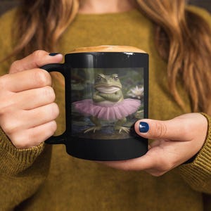May include: A black ceramic mug featuring a printed graphic of a green frog wearing a pink tutu with its arms crossed. The frog is positioned in a pond setting with water lilies visible in the background. The mug has a smooth finish and a handle.