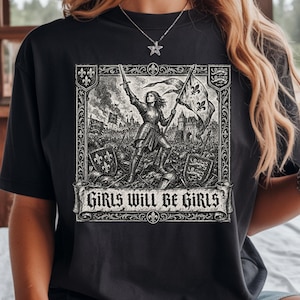 May include: A black short-sleeved t-shirt featuring a detailed monochromatic graphic print of a medieval warrior in armor holding a sword and flag, surrounded by fleur-de-lis symbols and the text "GIRLS WILL BE GIRLS" in a gothic font.