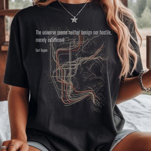 Op de afbeelding: Een zwart t-shirt met korte mouwen voorzien van een afbeelding van abstracte lijnen en de tekst: The universe seems neither benign nor hostile, merely indifferent. Carl Sagan. Het ontwerp bevat dunne witte en oranje lijnen die op een kaart lijken.