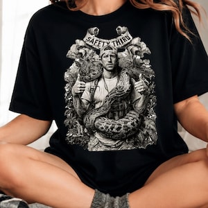 May include: A black short-sleeve t-shirt featuring a detailed monochromatic graphic of a person wearing a cap with a large snake coiled around their shoulders and torso. The text "SAFETY THIRD" is printed on a banner above the figure.