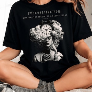 May include: A black t-shirt featuring a graphic print of a person with a cloud of smoke for hair, an alarm clock, and a smartphone. The text reads "PROCRASTINATION" and "WORKING TOMORROW FOR A BETTER TODAY" in white lettering above the central illustration.