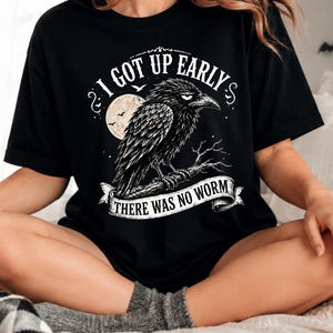 Könnte beinhalten: Ein schwarzes Kurzarm-T-Shirt mit einem weißen Grafikdruck einer Krähe, die auf einem Ast vor einem Vollmond sitzt. Der Text lautet I GOT UP EARLY und THERE WAS NO WORM, angeordnet in einem kreisförmigen Design mit dekorativen Schnörkeln.