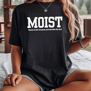 Może przedstawiać: Czarny t-shirt z krótkim rękawem z napisem MOIST dużymi białymi literami oraz mniejszym białym tekstem poniżej: Because at least one person you know hates this word. Koszulka posiada okrągły dekolt i charakteryzuje się luźnym krojem.