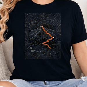 Puede incluir: Una camiseta negra de manga corta con un gráfico de mapa topográfico blanco en el pecho. El diseño incluye un sendero naranja, marcadores de picos de montaña blancos y etiquetas de texto como Base Camp, The Ridgeline Trail y Glacier Lake.