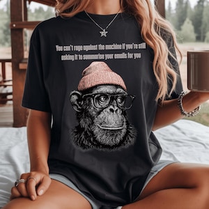 May include: A black t-shirt featuring a graphic of a chimpanzee wearing a pink beanie and black-rimmed glasses. Above the graphic, the text reads: You can't rage against the machine if you're still asking it to summarize your emails for you.