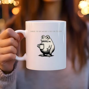 Può includere: Una tazza in ceramica bianca con un'illustrazione in bianco e nero di una rana che tiene in mano una tazzina. Sopra la rana, il testo recita: I HAVE TO SAY WEIRD STUFF OR I'LL DIE. La tazza presenta un manico standard e un design cilindrico.