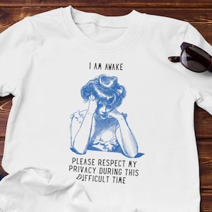 May include: White t-shirt with a blue illustration of a woman and the text "I AM AWAKE PLEASE RESPECT MY PRIVACY DURING THIS DIFFICULT TIME." Sunglasses and blue jeans are also visible.