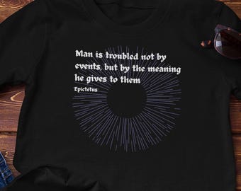 May include: Black t-shirt with the quote "Man is troubled not by events, but by the meaning he gives to them" and a circular design. The shirt is paired with blue jeans and sunglasses on a wooden surface.