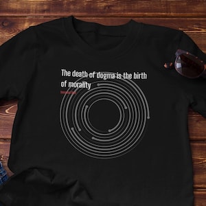 May include: Black t-shirt with the text "The death of dogma is the birth of morality" and a circular graphic. The shirt is paired with blue jeans and sunglasses. A small potted succulent is also visible.