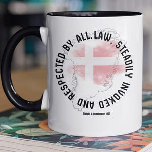 Può includere: Tazza in ceramica bianca con manico e bordo neri, con un disegno a mappa con una croce rossa e la scritta "RISPETTATO DA TUTTA LA LEGGE, COSTANTEMENTE INVOCATO E". La tazza è appoggiata su un libro.