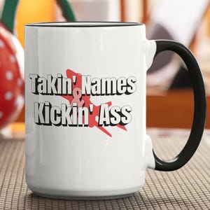 Pode incluir: Caneca de cerâmica branca com alça e borda pretas. A caneca apresenta o texto "Takin' Names Kickin' Ass" em letras pretas e vermelhas. Um gráfico de respingos vermelho também está incluído.