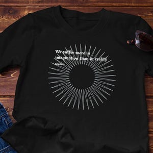 Puede incluir: Camiseta negra con texto blanco y un diseño circular. El texto dice: "Sufrimos más en la imaginación que en la realidad" de Séneca. La camiseta se combina con unos vaqueros azules y gafas de sol sobre una superficie de madera.