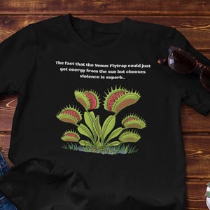 May include: Black t-shirt featuring a Venus Flytrap graphic with the text: "The fact that the Venus Flytrap could just get energy from the sun but chooses violence is superb..." Also includes blue jeans and sunglasses.
