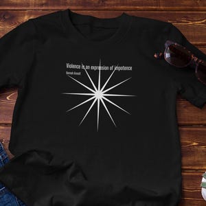 Op de afbeelding: Zwart T-shirt met een witte sterrenburst en de quote "Geweld is een uitdrukking van onmacht" van Susan Sontag. Het shirt wordt gecombineerd met een blauwe spijkerbroek en een zonnebril. Er is ook een kleine potplant aanwezig.