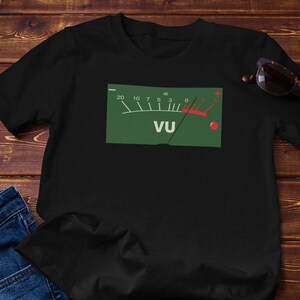 May include: Black t-shirt with a green graphic of a VU meter, featuring a red needle and the letters "VU". The shirt is paired with blue jeans and sunglasses on a wooden surface. A small succulent plant in a white pot is also present.