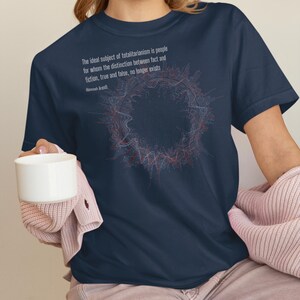 May include: A navy blue short-sleeved t-shirt featuring a circular abstract graphic in red and blue lines. The text reads: The ideal subject of totalitarianism is people for whom the distinction between fact and fiction, true and false, no longer exists. Hannah Arendt.