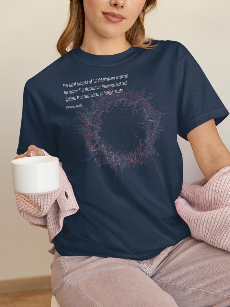 May include: A navy blue short-sleeved t-shirt featuring a circular abstract graphic in red and blue lines. The text reads: The ideal subject of totalitarianism is people for whom the distinction between fact and fiction, true and false, no longer exists. Hannah Arendt.
