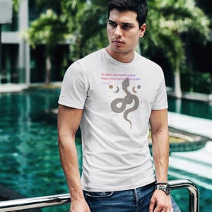 May include: White t-shirt with a gray snake graphic and text. The shirt features a crew neck and short sleeves. The text reads "The urge to save humanity is almost always a false front for the urge to rule it".