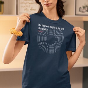 May include: Navy blue t-shirt with the text "The death of dogma is the birth of morality" and a circular graphic. The shirt is a crew neck style and is made of a soft material.