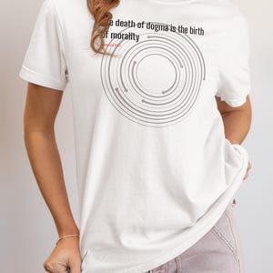 May include: White t-shirt with the text "The death of dogma is the birth of morality" and a gray circular design. The shirt is a short-sleeved, casual style.