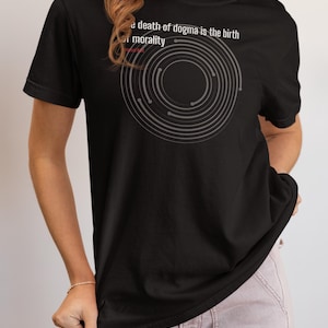 May include: Black t-shirt with the text "the death of dogma is the birth of morality" and a gray circular design. The shirt is a crew neck and has short sleeves. The model is wearing light pink pants.