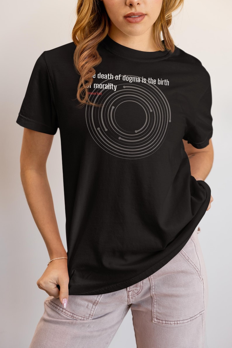May include: Black t-shirt with the text "the death of dogma is the birth of morality" and a gray circular design. The shirt is a crew neck and has short sleeves. The model is wearing light pink pants.