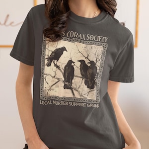 Op de afbeelding: Een antracietgrijs t-shirt met ronde hals en een vierkante grafische print van vier zwarte kraaien op kale boomtakken. De tekst boven de afbeelding luidt CORAX SOCIETY en de tekst eronder luidt LOCAL MURDER SUPPORT GROUP.