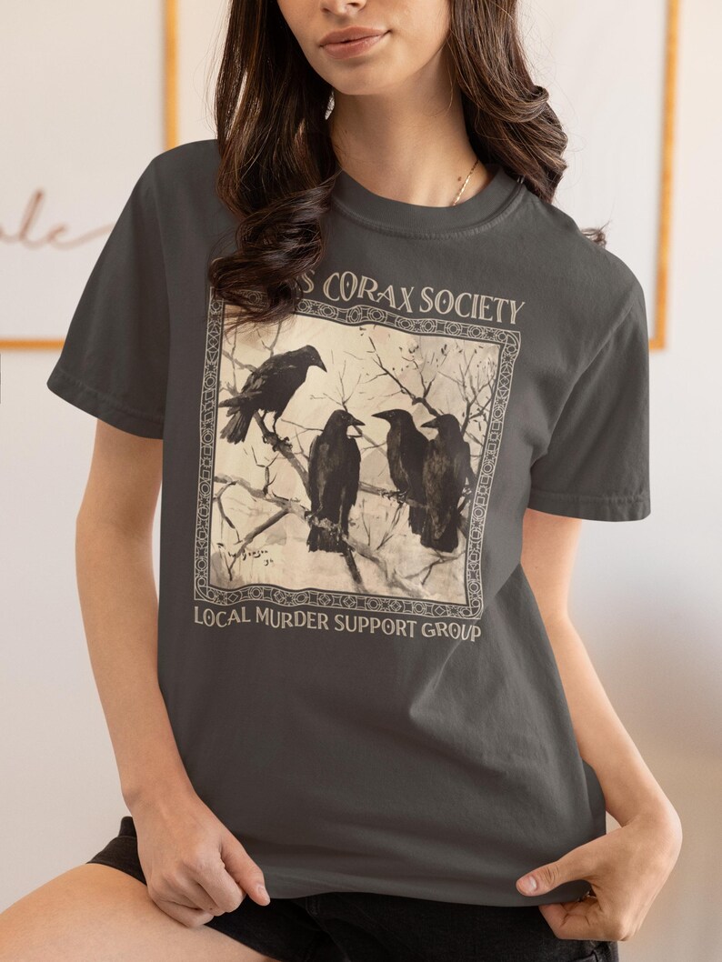 Op de afbeelding: Een antracietgrijs t-shirt met ronde hals en een vierkante grafische print van vier zwarte kraaien op kale boomtakken. De tekst boven de afbeelding luidt CORAX SOCIETY en de tekst eronder luidt LOCAL MURDER SUPPORT GROUP.