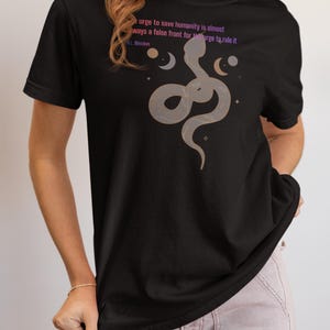 May include: Black t-shirt featuring a stylized snake design with moons and stars. The shirt includes the quote "urge to save humanity is almost always a false front for the urge to rule it" by H.L. Mencken.