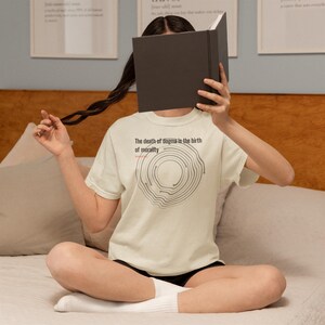 May include: Cream-colored t-shirt with the text "The death of dogma is the birth of morality" and a gray circular design. The shirt is worn by a person holding a book.