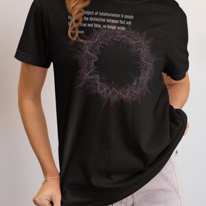 May include: A black short-sleeve t-shirt featuring a circular abstract graphic in red and blue lines. The text reads: The ideal subject of totalitarianism is people for whom the distinction between fact and fiction, true and false, no longer exists. Hannah Arendt.