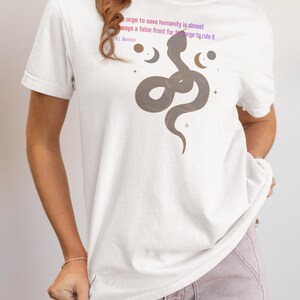 May include: White t-shirt featuring a brown snake illustration with crescent moons and stars. The shirt has text that reads "urge to save humanity is almost ways a false front for urge to rule it" and "H.L. Mencken".