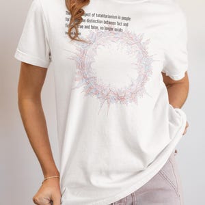 May include: A white short-sleeved t-shirt featuring a circular graphic composed of red and blue lines. Above the graphic, black text reads: The ideal subject of totalitarianism is people for whom the distinction between fact and fiction, true and false, no longer exists. Hannah Arendt.